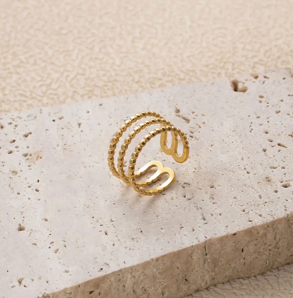 A gold-plated triple-band spiral ring with textured and smooth segments, displayed on a light beige stone slab against a textured beige background.