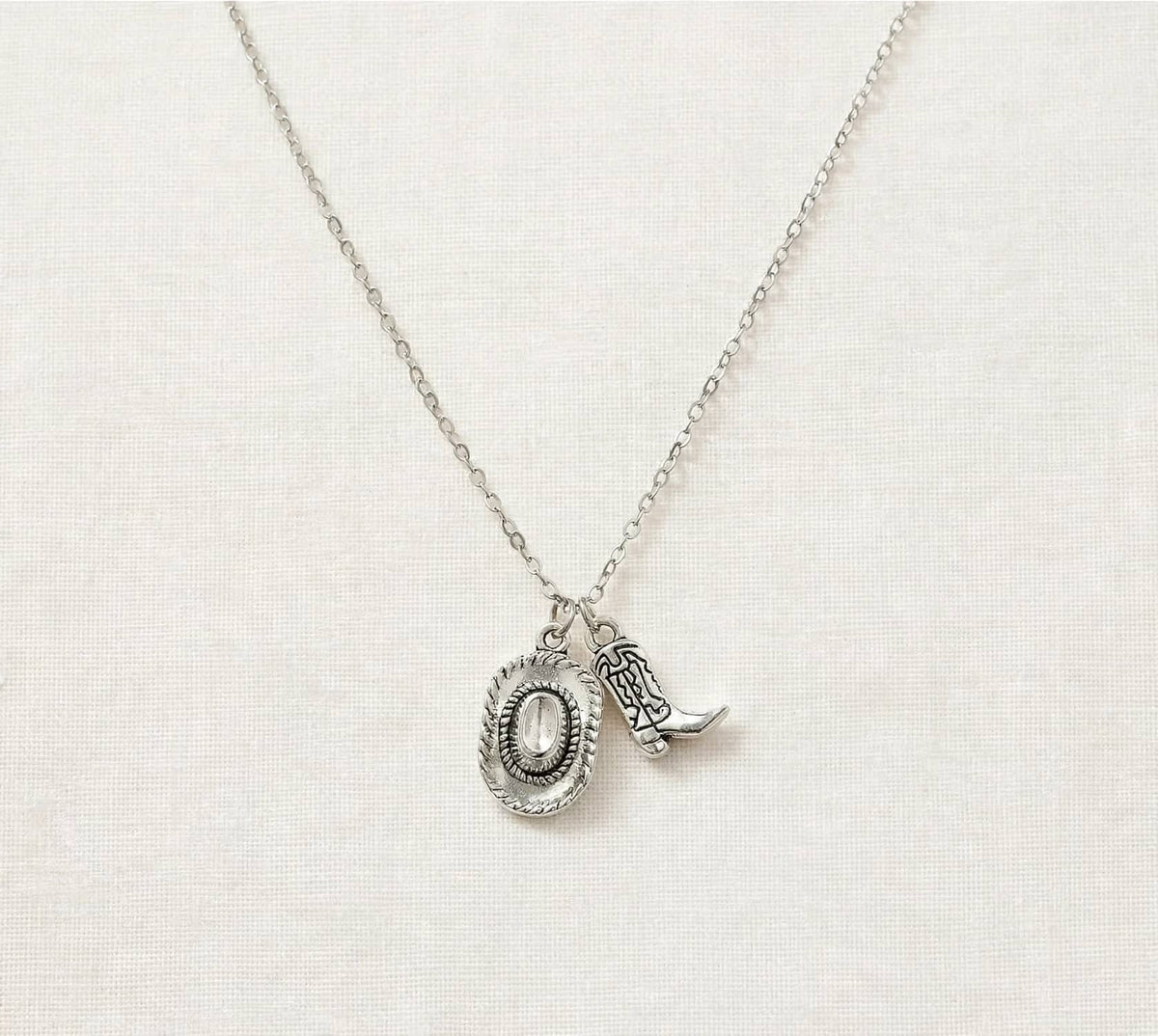 A silver necklace with a textured oval pendant and a small cowboy boot charm, displayed on a neutral cream-colored fabric background.