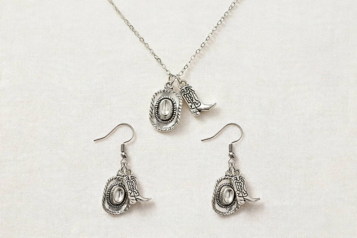 Silver-toned jewelry set featuring a necklace and matching drop earrings with cowboy boot and oval medallion charms, displayed on a textured off-white fabric background.