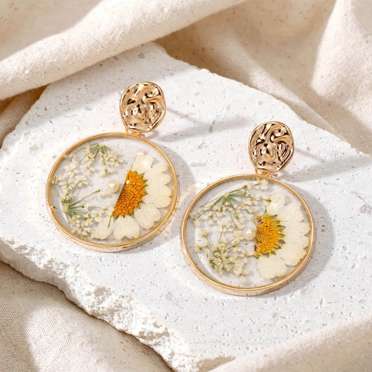 A pair of gold-toned dangle earrings with round clear resin domes containing dried white daisies and baby's breath, displayed on a textured white stone slab with beige fabric in the background.
