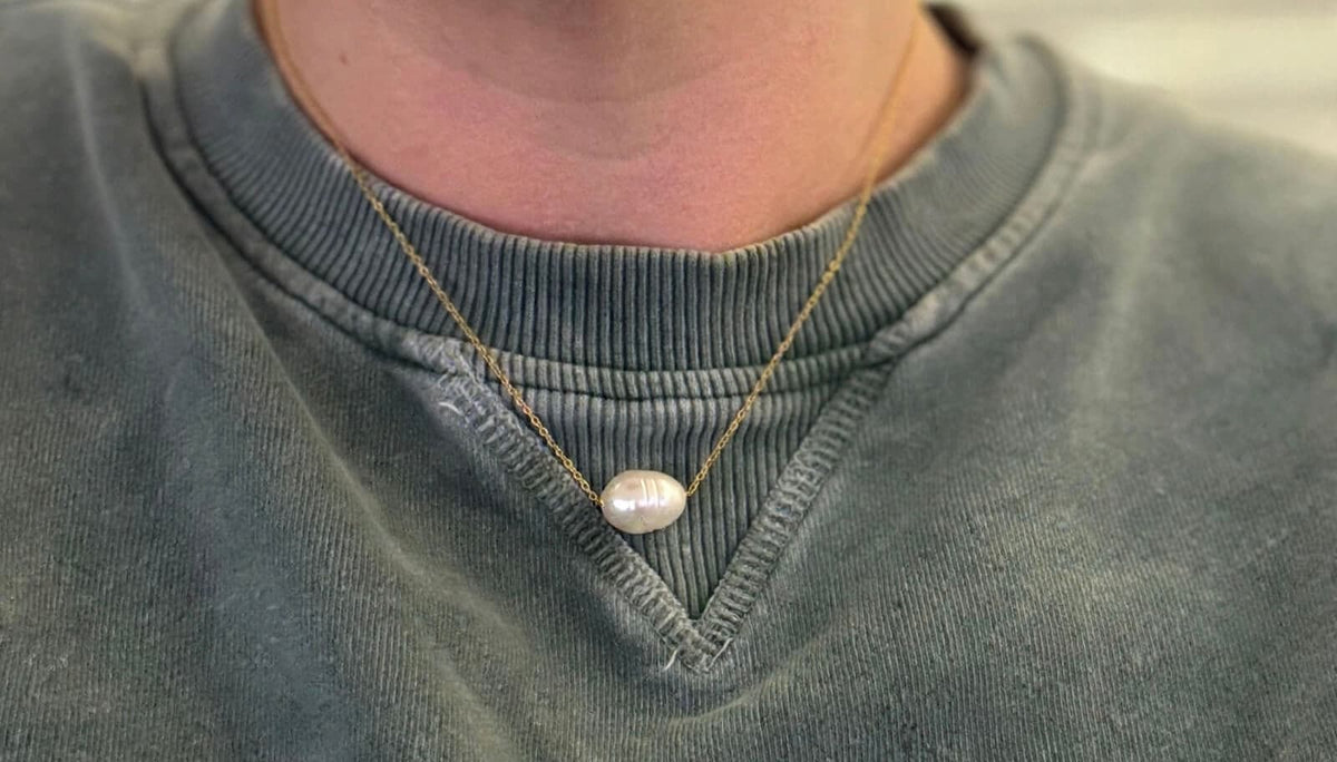 Close-up of a gold-colored chain necklace with a white freshwater pearl pendant worn over a green sweatshirt.