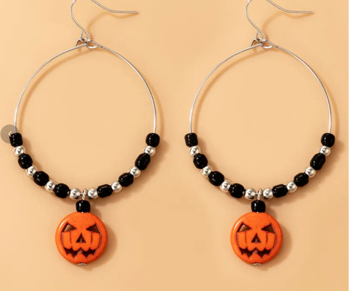 A pair of silver-tone hoop earrings with black and silver beads, each hanging from a pumpkin-shaped orange charm, displayed against a solid peach background.