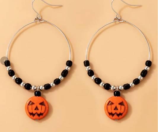 A pair of silver-tone hoop earrings with black and silver beads, each hanging from a pumpkin-shaped orange charm, displayed against a solid peach background.
