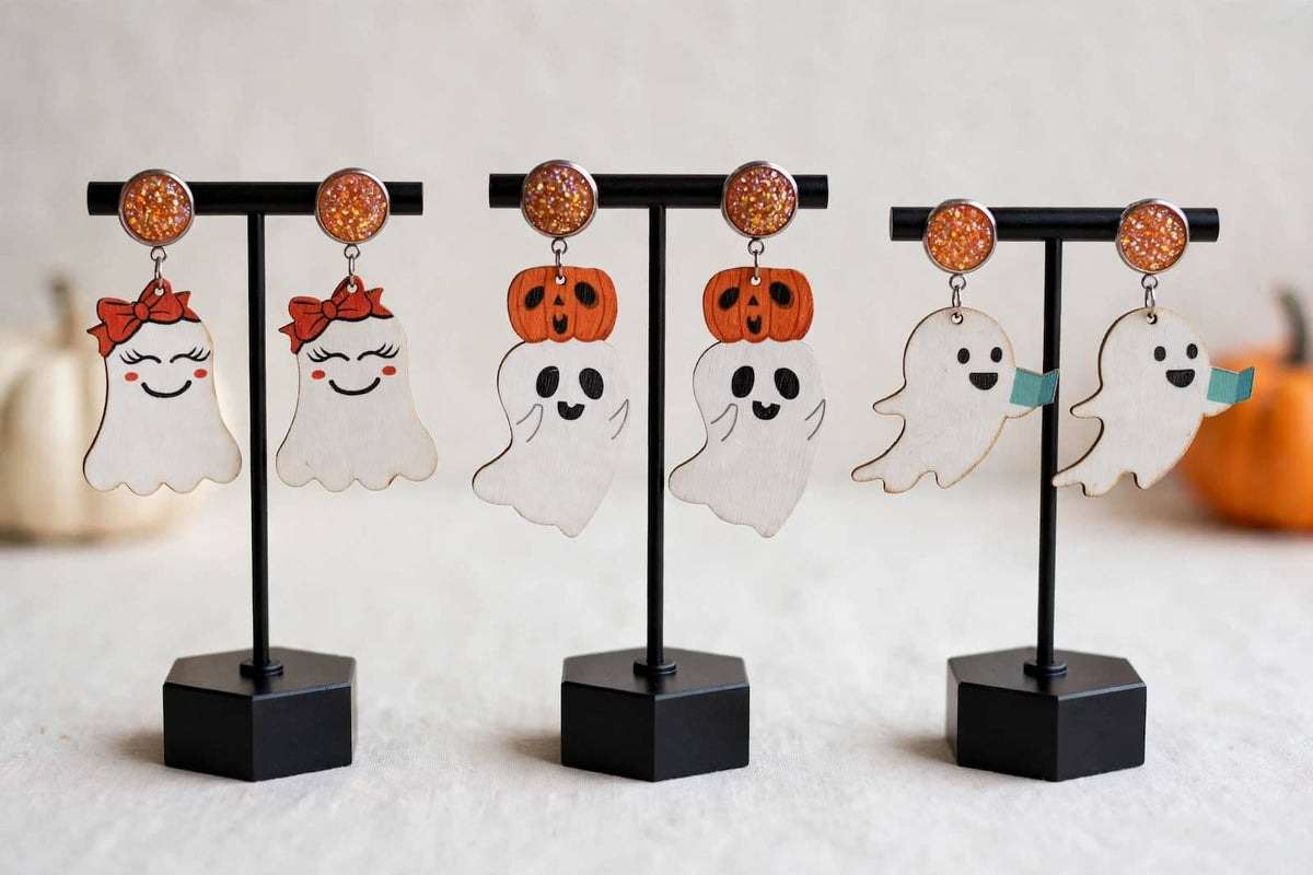 Four pairs of Halloween-themed dangling earrings displayed on black T-stands. The earrings feature cute ghost and pumpkin designs with orange glittery studs, shown against a soft, light beige background with blurred pumpkins.