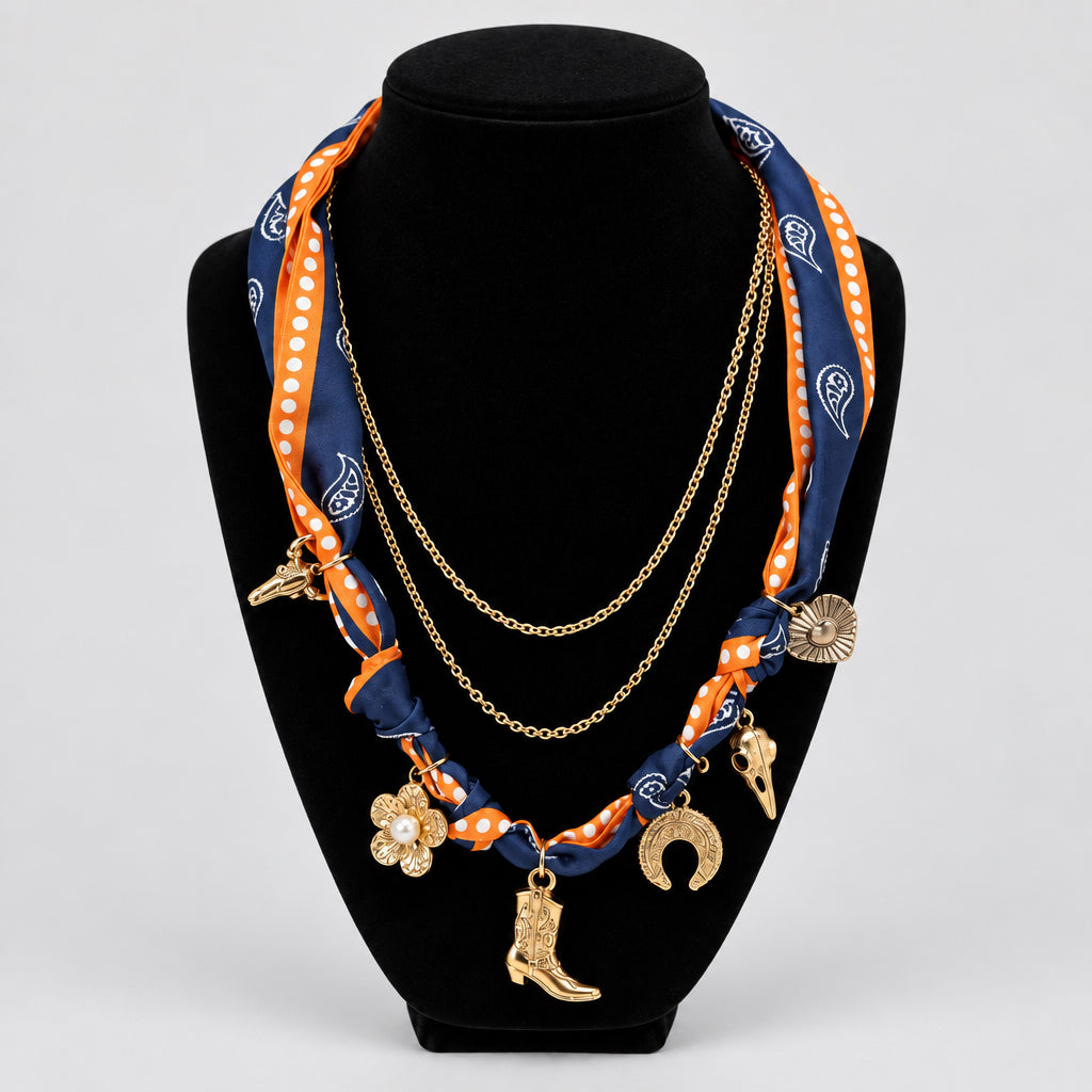 A double-layered necklace displayed on a black velvet jewelry bust. The necklace features a navy blue and orange paisley silk scarf wrapped with gold polka dots, layered over a gold chain, adorned with western-themed charms including a cowboy boot, horseshoe, and clover.