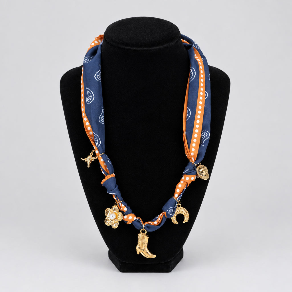A western-themed silk scarf styled as a necklace on a black velvet display bust. The scarf features a navy blue base with orange paisley patterns, orange polka-dot borders, and gold-toned charms shaped like a cowboy boot, a flower, and a crescent moon.