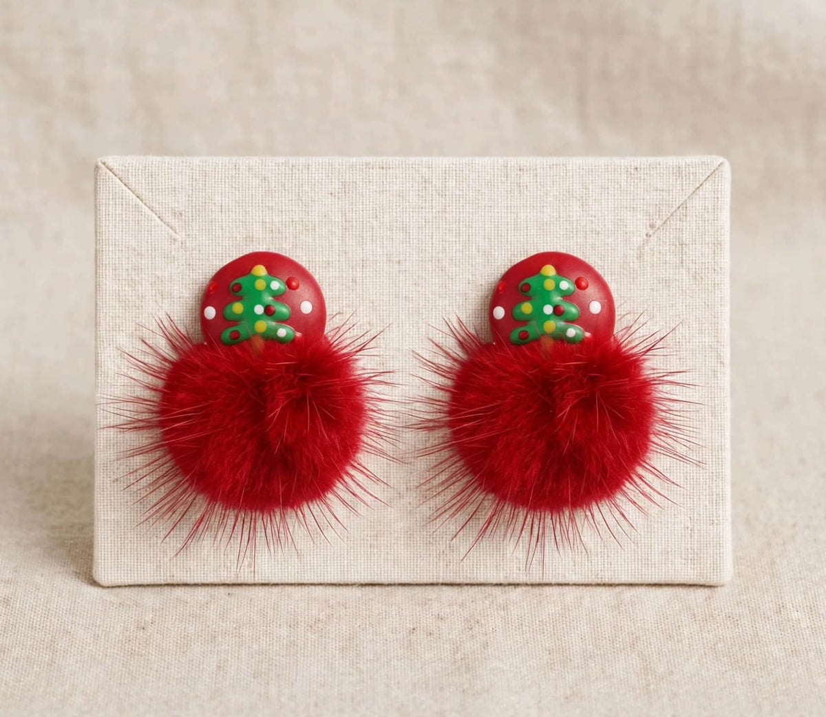A pair of festive red pom-pom earrings with a small red Christmas tree charm on top, displayed on a square beige fabric card against a neutral beige background.