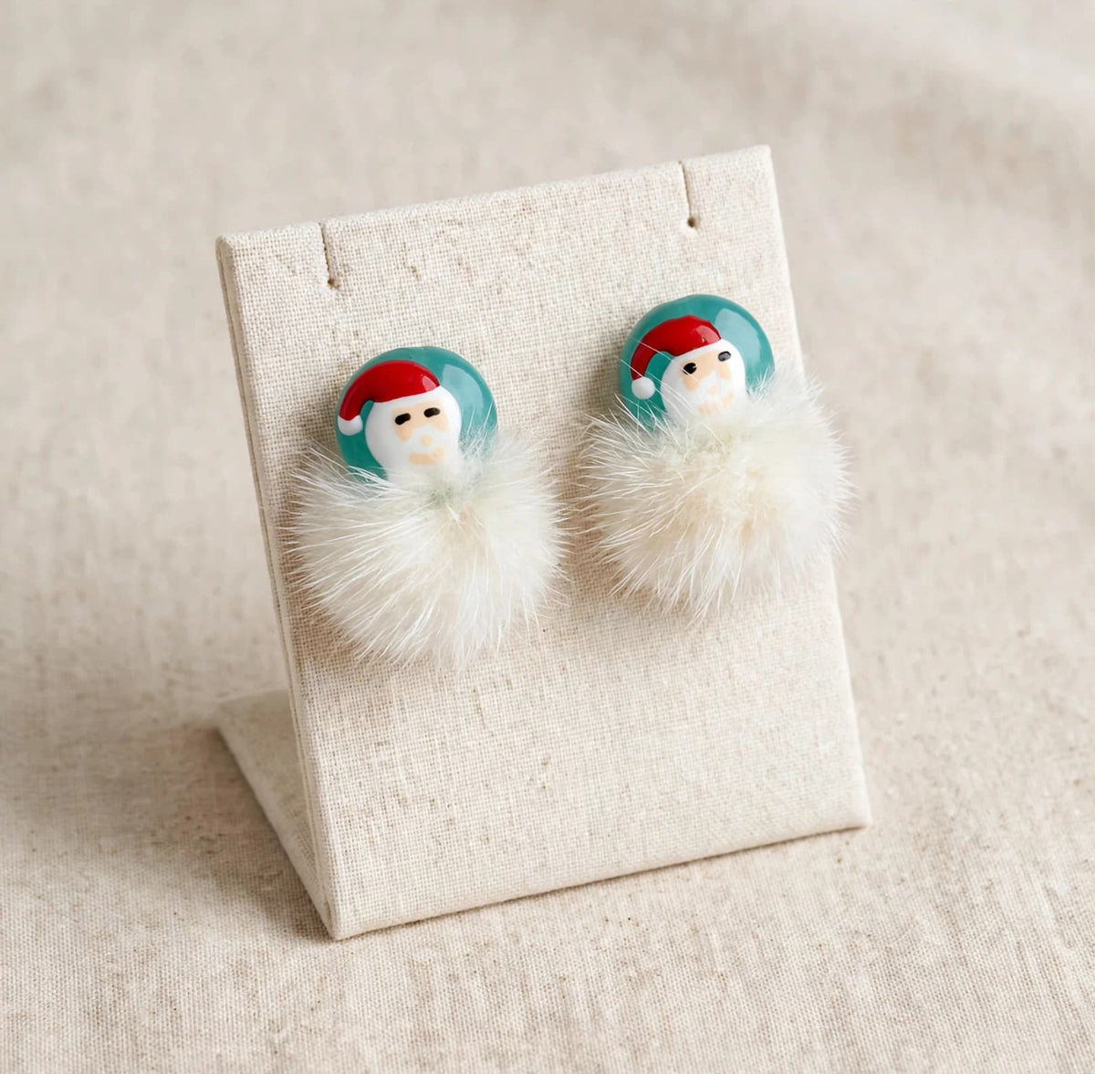 A pair of holiday-themed dangle earrings displayed on a beige fabric stand. Each earring features a round turquoise blue top with a white Santa face wearing a red hat, hanging from a fluffy cream faux-fur pom-pom.